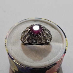 May include: A silver ring with a large, round, deep red gemstone at the center, surrounded by smaller clear stones. The ring is displayed in a small, round box with a floral design.