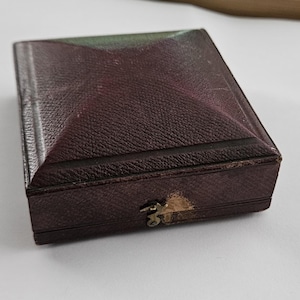 May include: A rectangular, burgundy-colored box with a textured surface and a pyramidal lid. The box has a small, tarnished metal clasp. The box is likely for jewelry or other small items.