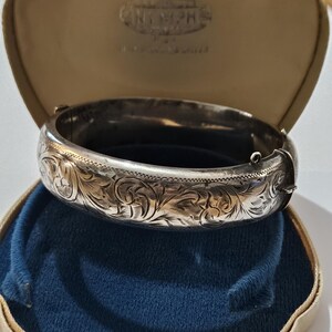 Peut inclure: Un bracelet jonc en argent avec des gravures florales complexes, présenté dans un écrin vintage ouvert, en forme de coquillage, avec un intérieur en velours bleu. L'étui est beige et porte le mot "NYMPHS".