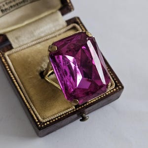 Puede incluir: Un anillo con una gran piedra preciosa rectangular de color magenta facetada, montada en una banda de metal dorado. El anillo se presenta dentro de un joyero marrón oscuro con bisagras, forro de terciopelo color crema y borde dorado.