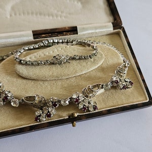 Peut inclure: Un ensemble collier et bracelet en strass vintage dans une boîte en velours marron. Le collier présente un design délicat avec des pierres rouges et claires. Le bracelet est un modèle simple à une rangée.