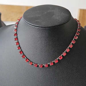 Könnte beinhalten: Halskette mit roten, quadratisch geschliffenen Edelsteinen, präsentiert auf einer schwarzen Schaufensterpuppe. Die silberne Kette trägt gleichmäßig verteilte, leuchtend rote Steine. Die Schaufensterpuppe steht vor einem neutralen Hintergrund, der die Farbe und das Design des Schmucks hervorhebt.