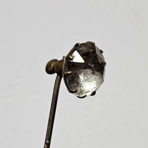 Puede incluir: Un broche de sombrero vintage con una gema de cristal transparente y facetada. La gema está engastada en un engaste de metal color latón y se asienta sobre un vástago de metal retorcido.