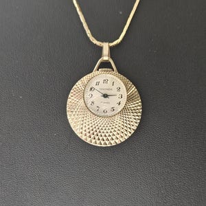 Puede incluir: Un reloj colgante Sekonda dorado con una esfera circular texturizada. El reloj presenta una esfera blanca con números y agujas negras, suspendida de una delicada cadena dorada. La esfera del reloj mide aproximadamente 2,5 cm de diámetro.