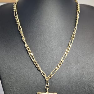 Op de afbeelding: Een goudkleurige ketting met een Figaro-kettingontwerp en een T-bar hanger. De ketting wordt getoond op een zwarte mannequin. De schakels van de ketting wisselen af tussen langwerpige en ovale vormen, wat een klassieke en elegante uitstraling creëert.