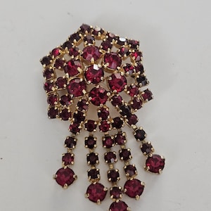 May include: A vintage brooch with a gold-tone setting, adorned with numerous round and oval red gemstones. The brooch features a cluster of stones at the top, with cascading dangles of gems. A classic piece of jewelry.