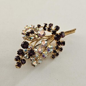 Puede incluir: Broche vintage dorado con diseño floral. El broche presenta grupos de cristales morados oscuros e iridiscentes, dispuestos para parecer hojas y flores. Un accesorio clásico.