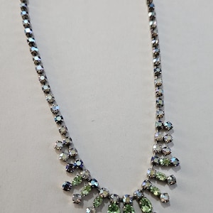 May include: A vintage necklace with a silver-tone chain and iridescent rhinestones. The necklace features a central cluster of green and clear stones, with smaller clusters of stones cascading down. The necklace is designed to be worn around the neck.