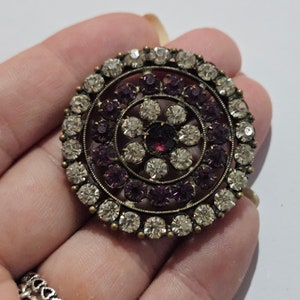 Op de afbeelding: Een ronde, zilverkleurige broche met een cirkelvormig ontwerp van heldere en paarse strass.