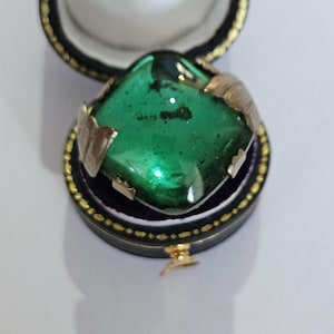 Peut inclure: Une pierre précieuse carrée vert émeraude sertie dans un métal doré, présentée dans un écrin noir ouvert avec des bordures dorées. L'écrin a un intérieur violet et est posé sur un fond blanc.