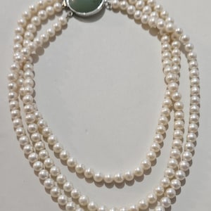 Puede incluir: Collar de perlas de tres hilos con un cierre ovalado de color jade. Las perlas blancas y redondas están dispuestas en forma circular.