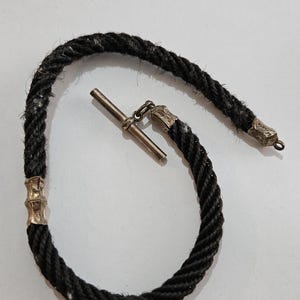 May include: A black rope bracelet with silver-toned metal accents. The bracelet features a twisted rope design with decorative metal ends and a T-bar clasp. The bracelet is likely vintage or antique.