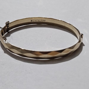 May include: A gold-coloured bangle bracelet with a geometric, diamond-shaped pattern. The bracelet has a clasp closure and a safety chain. The text "BCI METAL CORP" is stamped on the inside. Jewellery.