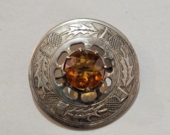 May include: A silver-toned brooch with an amber-colored gemstone center. The brooch features a circular design with etched thistle motifs around the edge. The gemstone is round and faceted, adding sparkle to the piece.