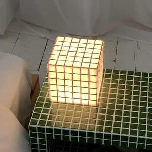 以下が含まれることがあります： 緑色のタイル製のナイトスタンドに棚と白い立方体のランプが置かれています。ランプは点灯しており、温かい光を放っています。