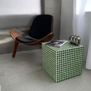 Puede incluir: Una mesa auxiliar de cubo de azulejos verdes con una revista encima y jarrones plateados a un lado. La mesa está delante de una silla de cuero negro con marco de madera.