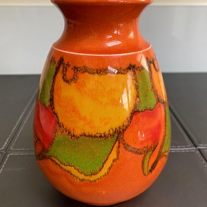 Op de afbeelding: Een keramische vaas met een bolvormige body en een smalle hals. De vaas is voornamelijk oranje met abstracte ontwerpen in rood, groen en geel, omlijnd in zwart. De rand is wit.