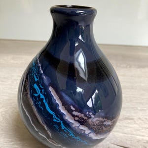 May include: A dark blue ceramic vase with a glossy finish. The vase has a rounded body and a narrow neck. The design features a blend of blue, black, and white abstract patterns. The vase is approximately 15 cm tall.