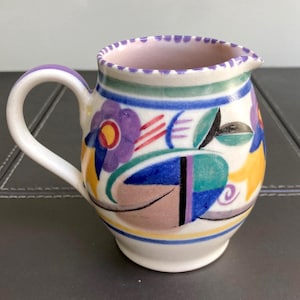 Könnte beinhalten: Ein Sahnekännchen aus Keramik mit handgemaltem Design aus violetten Blumen, gelben Akzenten und türkisfarbenen geometrischen Formen auf weißem Grund. Der Rand ist mit einem violetten Punktmuster verziert und hat einen geschwungenen Henkel.
