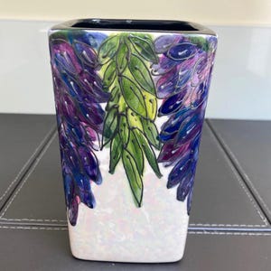 Puede incluir: Un jarrón de cerámica de forma cuadrada con un acabado blanco iridiscente brillante. El exterior presenta un diseño pintado a mano de flores de glicinia moradas colgantes y hojas verdes vibrantes, con un borde negro sólido en la abertura.