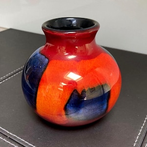 May include: A small, round, bulbous vase with a glossy finish. The exterior features a vibrant color palette of bright orange and deep red, accented by large, dark blue patches that wrap around the sides. The rim is a solid, smooth black.