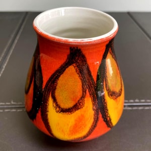 May include: A small ceramic vase with a rounded body and a wide opening. The exterior features a vibrant orange and yellow glaze decorated with bold, dark brown hand-painted circular patterns. The interior of the vase is finished in white.