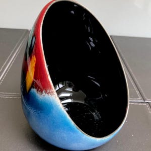 Puede incluir: Un cuenco de cerámica con una forma asimétrica única. El exterior presenta una vibrante paleta de colores, que va del azul en la base al rojo y amarillo en la parte superior. El interior es negro brillante.