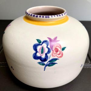 May include: A round, off-white ceramic vase featuring a hand-painted floral motif in shades of blue, pink, purple, and green. The rim is decorated with a purple dotted pattern and a solid yellow band, while the interior is a soft pink hue.