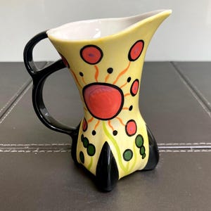 May include: A ceramic pitcher with a yellow base, featuring a black handle and black base accents. The exterior is decorated with a large red circular motif surrounded by smaller red and green dots, along with orange radiating lines on a yellow field.