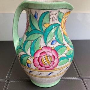 Może przedstawiać: Ceramiczny dzbanek z ręcznie malowanym wzorem kwiatowym w żywych kolorach różowym, żółtym i zielonym. Wzór zawiera stylizowane liście i kwiaty na jasnym tle, z zielonym uchwytem oraz żółtym paskiem w dolnej części naczynia.