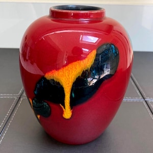 Puede incluir: Un jarrón de cerámica bulboso con un acabado rojo brillante, que presenta un patrón abstracto de goteo en esmalte negro y amarillo vibrante. El jarrón tiene un cuello corto y estrecho con un borde interior oscuro y una superficie lisa.