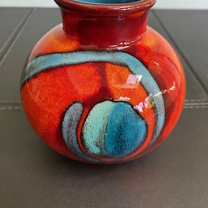 May include: A small, round ceramic vase with a glossy finish. The exterior features a vibrant orange and red base colour with a prominent, swirling light blue and dark grey abstract pattern. The interior of the vase is a solid, smooth light blue.