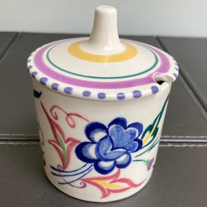 May include: A white ceramic sugar bowl with a lid, featuring a blue floral design and colourful accents. The lid has a handle and is decorated with yellow, purple, and blue stripes. The bowl has a spoon rest.