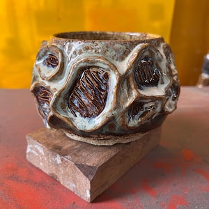 May include: A round ceramic bowl with a textured surface featuring raised, organic shapes. The exterior displays a mix of deep brown, glossy white, and muted green glazes, with dark brown recessed areas showing vertical, etched line patterns.