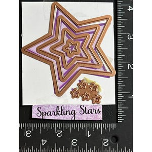 May include: A set of star-shaped metal die cuts in varying sizes, with a copper finish and purple backing. The image includes a small cluster of star cutouts and a label that reads "Sparkling Stars".