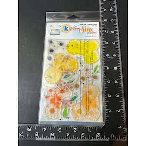 May include: A package of clear photopolymer stamps labeled Kitchen Sink Stamps Fresh as a Daisy, featuring various daisy flower and leaf designs. A black ruler is positioned along the side and bottom, showing the package is 4 inches wide.