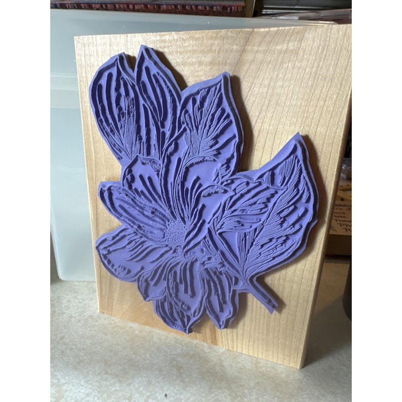 May include: A purple floral stamp with intricate details of petals and leaves is mounted on a rectangular wooden block. The stamp is a vibrant shade of purple, and the wood has a natural, light color. This stamp is ideal for crafting and scrapbooking.