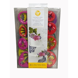 May include: A clear plastic package containing a Wilton Gum Paste Flower Cut-Outs Set. The set includes various colorful plastic cutters in red, yellow, pink, and purple shapes. The packaging features a cake decorated with gum paste flowers.