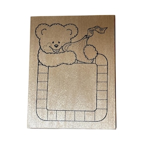 May include: A wooden stamp featuring a smiling teddy bear holding a gift bag with heart accents. The bear is positioned above a rectangular frame with a grid-like border, perfect for crafting and scrapbooking projects.