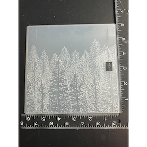 May include: A clear, rectangular embossing folder featuring a detailed forest scene with numerous evergreen trees. The trees are densely packed, creating a textured effect. The folder has a small, square logo in the corner.