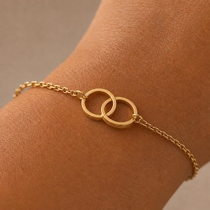 May include: A gold-toned chain bracelet featuring two interlocking circular rings as a central focal point. Each ring is engraved with a name, specifically 'Julie' on one and 'Kenneth' on the other, connected to a delicate link chain.