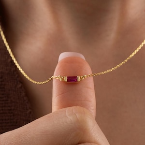 May include: A delicate gold necklace featuring a small, rectangular ruby pendant. The pendant is a vibrant red color and is set in a gold bezel. The necklace is shown against a brown background.