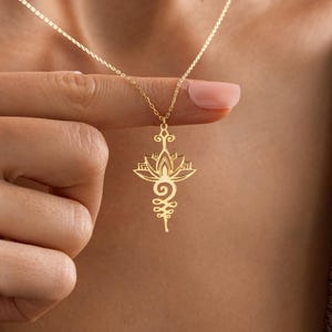 May include: A gold-colored necklace with a lotus flower pendant. The pendant features intricate cut-out details and a delicate chain. The necklace is being held up by a person's hand.
