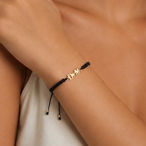 Könnte beinhalten: Ein schwarzes Armband aus Kordel mit zwei goldfarbenen Metallbuchstaben, D und M, getrennt durch ein kleines goldfarbenes Herzsymbol. Das Armband hat einen verstellbaren Schiebeknotenverschluss mit zwei kleinen goldfarbenen Perlen an den Enden.
