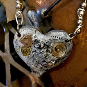 May include: A steampunk-style heart pendant necklace. The heart is textured with a gray and white finish, adorned with small clockwork gears and metal accents. The necklace has a silver chain and a black cord.