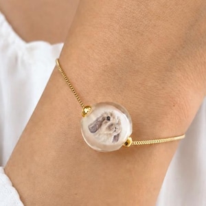 May include: A gold-toned chain bracelet featuring a central clear, spherical glass bead. Inside the bead is a detailed illustration of a brown lop-eared rabbit. The bracelet is secured with small gold-colored metal caps on either side of the bead.