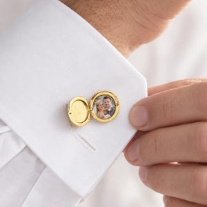 Peut inclure: Un bouton de manchette médaillon circulaire doré fixé sur une manchette de chemise blanche. Le médaillon est ouvert, révélant une petite photographie circulaire d'un couple à l'intérieur. Le métal a une finition polie et la charnière est visible.