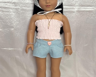 May include: A doll with long dark hair wearing a white lace headscarf, a light pink smocked tube top, a gold-tone necklace, light blue denim shorts with a pink heart-shaped button, beaded bracelets, white socks, and white sneakers with pink laces.