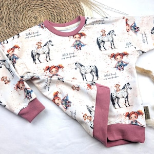 Puede incluir: Una sudadera blanca de manga larga con ribetes rosas. La sudadera tiene un patr&oacute;n repetido de una ni&ntilde;a con cabello rojo montando un caballo manchado. El texto "Wild. Frech und wunderbar" se repite en todo el patr&oacute;n.