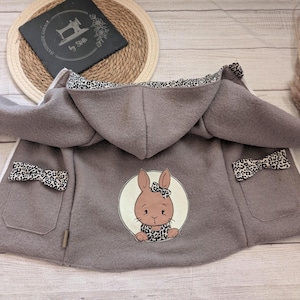 Könnte beinhalten: Eine taupefarbene Kapuzenjacke mit weißem Reißverschluss, langen Ärmeln mit hellbeigen Bündchen und zwei Taschen, die mit Leopardenmuster-Schleifen verziert sind. Ein kreisförmiges Motiv auf dem Rücken zeigt einen braunen Hasen mit Leoparden-Schleife.